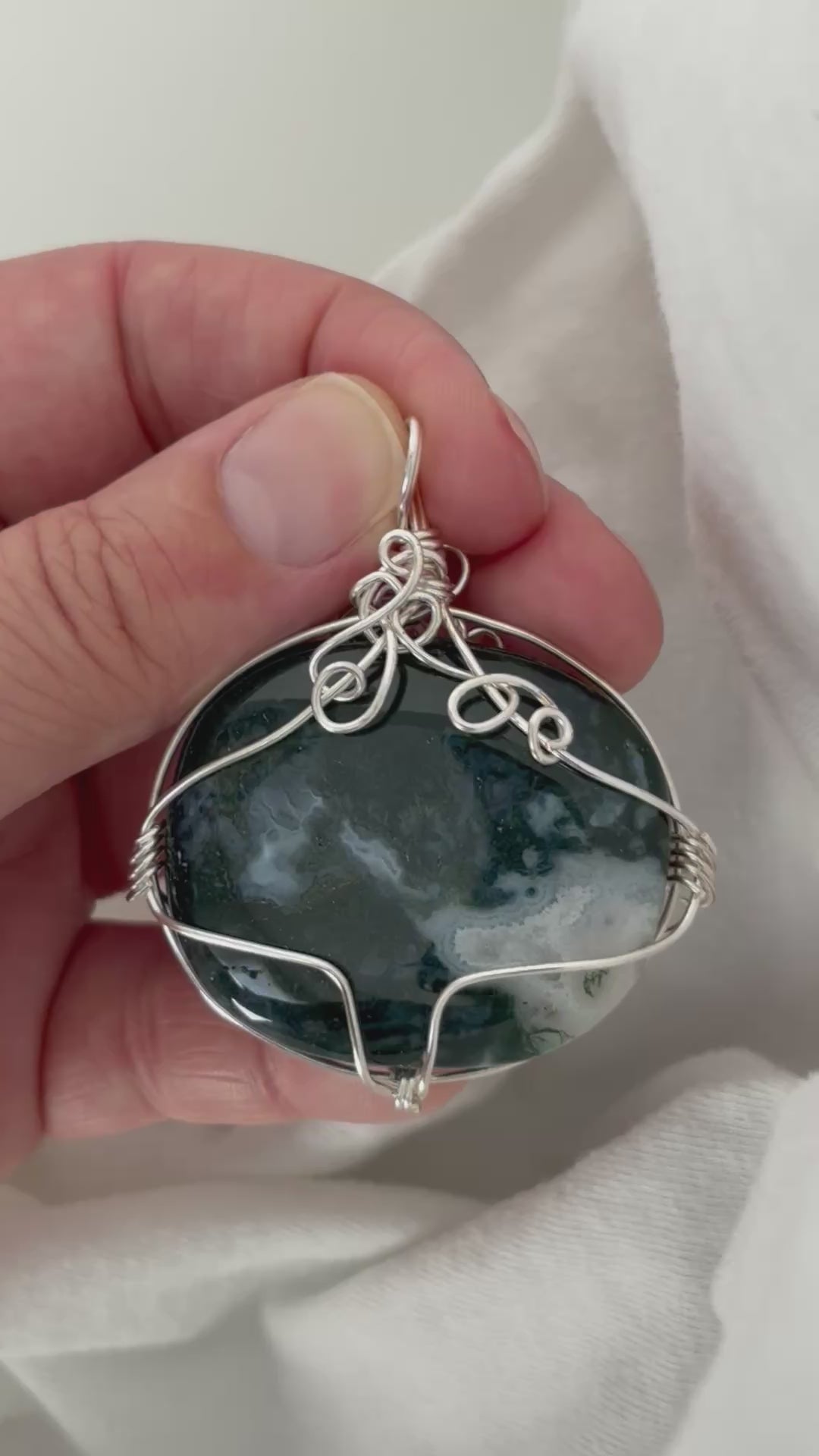 hand holding and moving the silver wire wrapped moss agate pendant around so all the sides of it are shown. done against a white background.