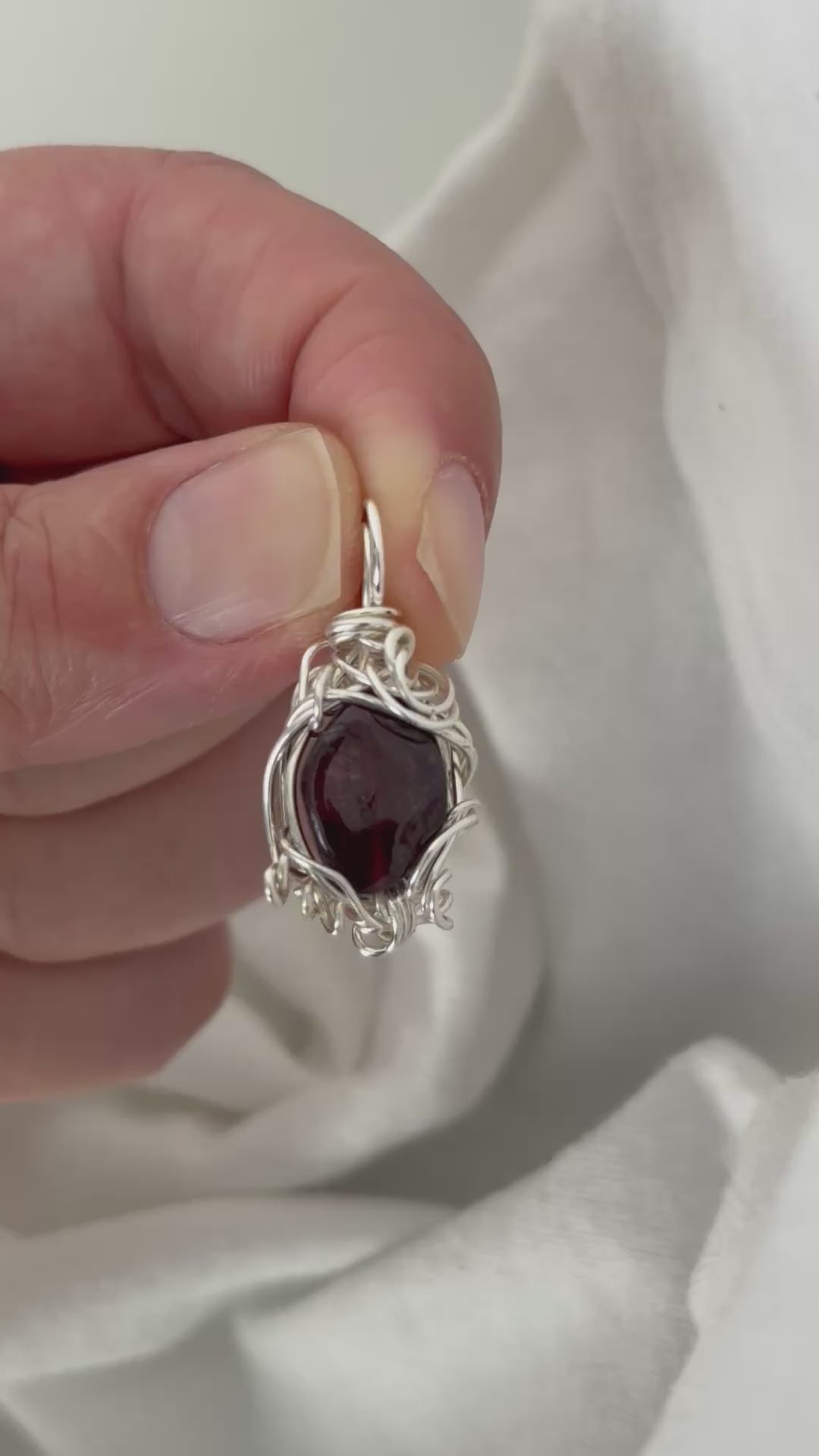 hand holding and moving the silver wire wrapped  garnet pendant around so all the sides of it are shown. done against a white background.