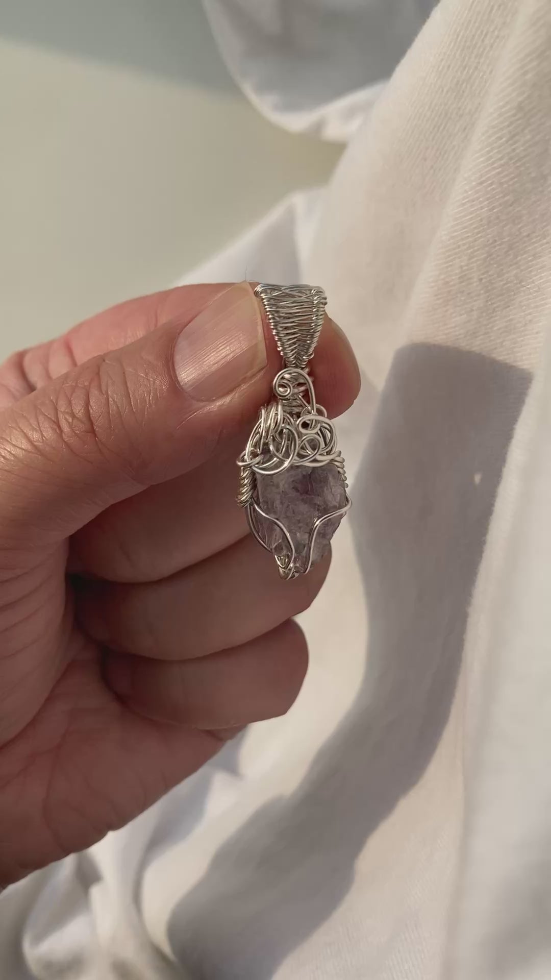 Lepidolite Pendant - Video of a wire-wrapped purple lepidolite stone pendant held between fingers over a white background, showing all sides of the pendant in natural lighting.