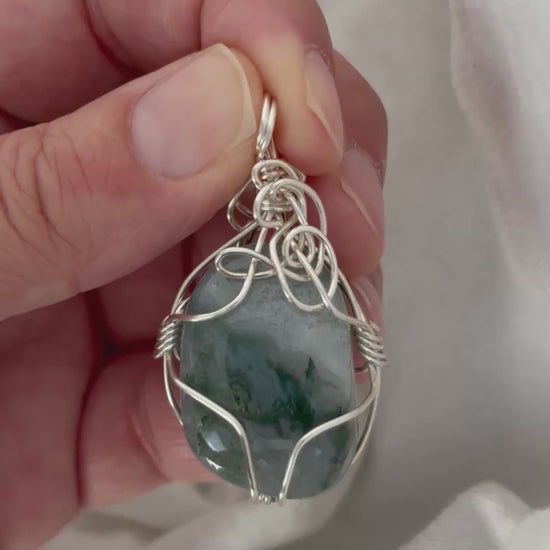 hand holding and moving the silver wire wrapped moss agate pendant around so all the sides of it are shown. done against a white background.