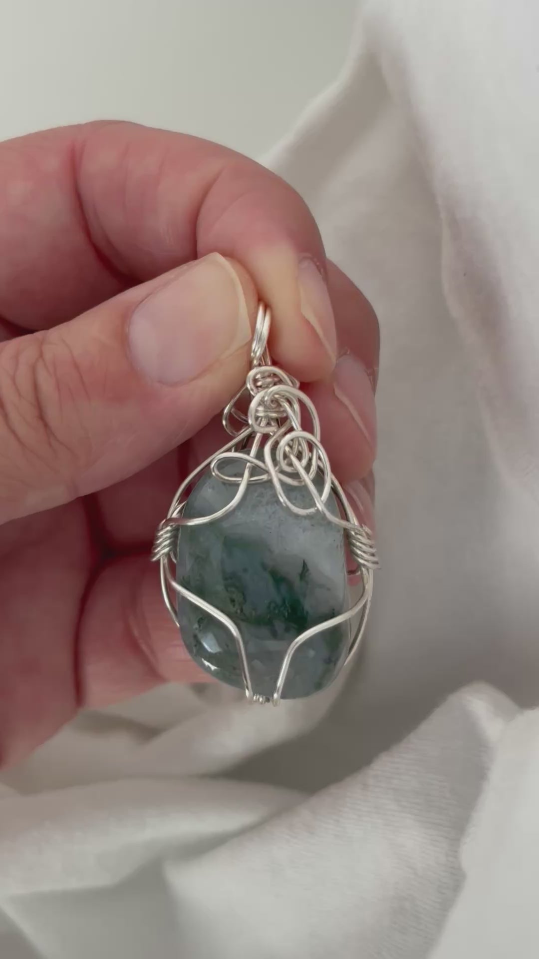 hand holding and moving the silver wire wrapped moss agate pendant around so all the sides of it are shown. done against a white background.