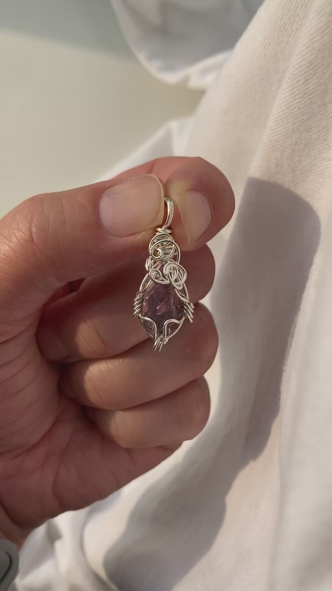Lepidolite Pendant - Video of a wire-wrapped purple lepidolite stone pendant held between fingers over a white background, showing both front and back sides in natural lighting.