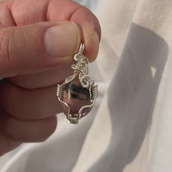 Rhodonite Pendant - Video of a wire-wrapped pink and black rhodonite stone pendant held between fingers over a white background, showing all sides of the pendant in natural lighting.