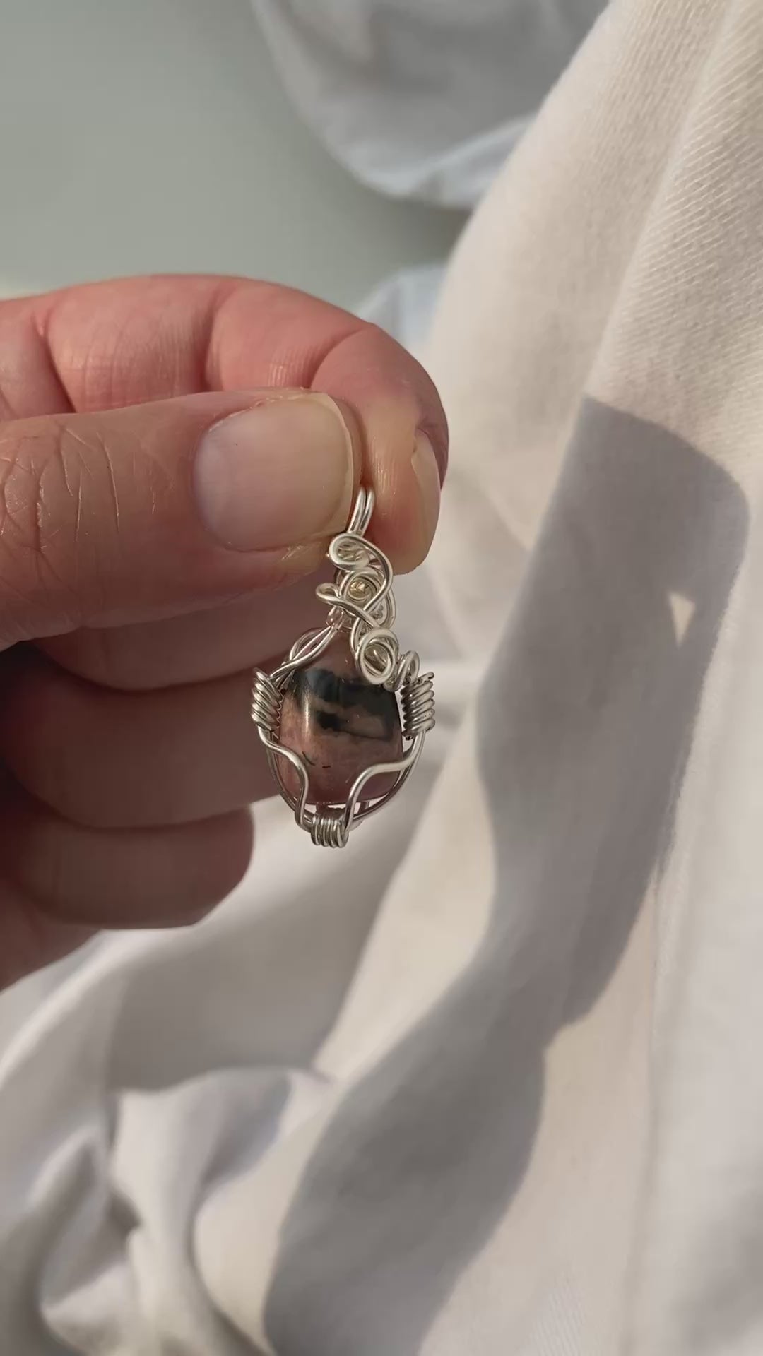 Rhodonite Pendant - Video of a wire-wrapped pink and black rhodonite stone pendant held between fingers over a white background, showing all sides of the pendant in natural lighting.
