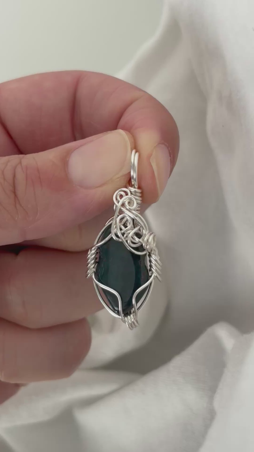 hand holding and moving the silver wire wrapped  bloodstone pendant around so all the sides of it are shown. done against a white background.