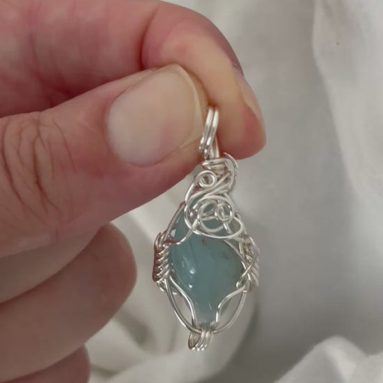 hand holding and moving the silver wire wrapped  aquamarine pendant around so all the sides of it are shown. done against a white background.
