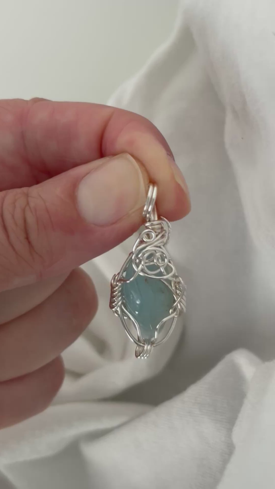 hand holding and moving the silver wire wrapped  aquamarine pendant around so all the sides of it are shown. done against a white background.
