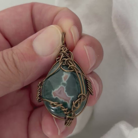 hand holding and moving the silver wire wrapped ocean jasper pendant around so all the sides of it are shown. done against a white background.