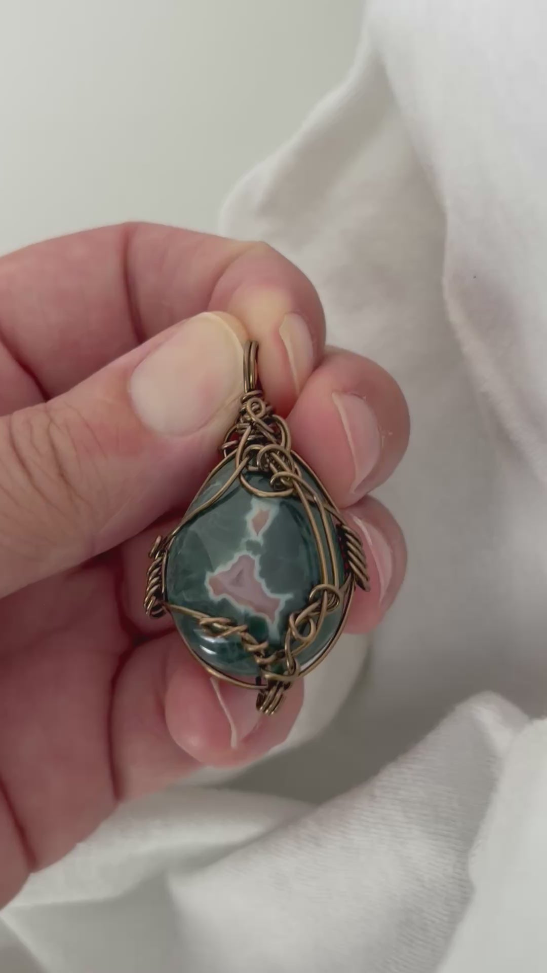 hand holding and moving the silver wire wrapped ocean jasper pendant around so all the sides of it are shown. done against a white background.