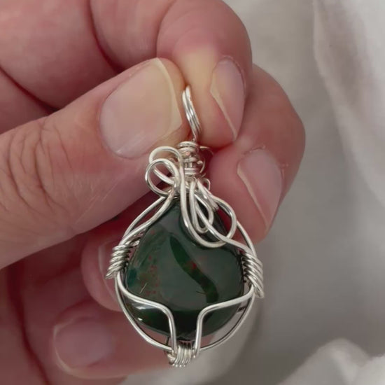 hand holding and moving the silver wire wrapped  bloodstone pendant around so all the sides of it are shown. done against a white background.