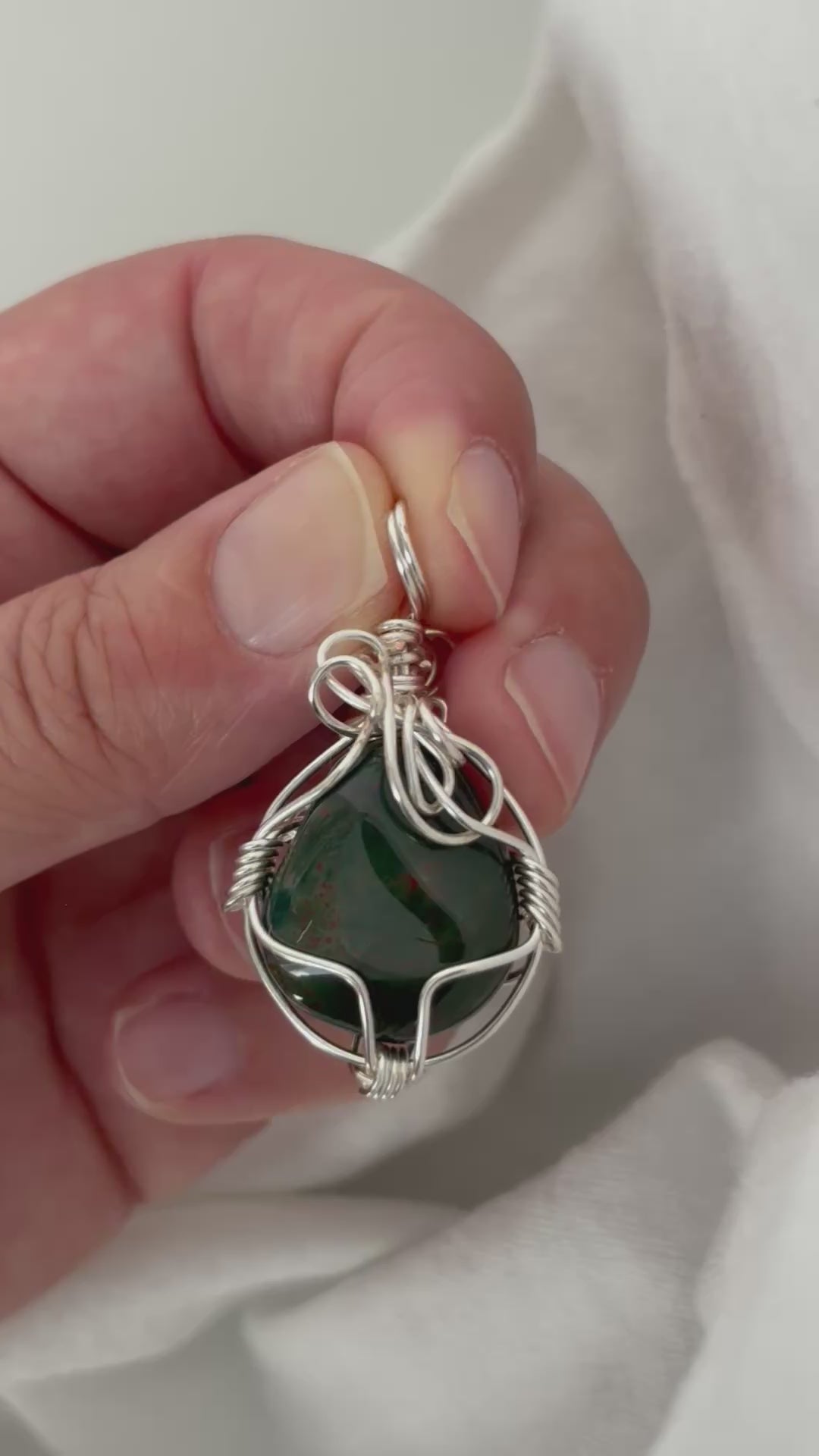 hand holding and moving the silver wire wrapped  bloodstone pendant around so all the sides of it are shown. done against a white background.