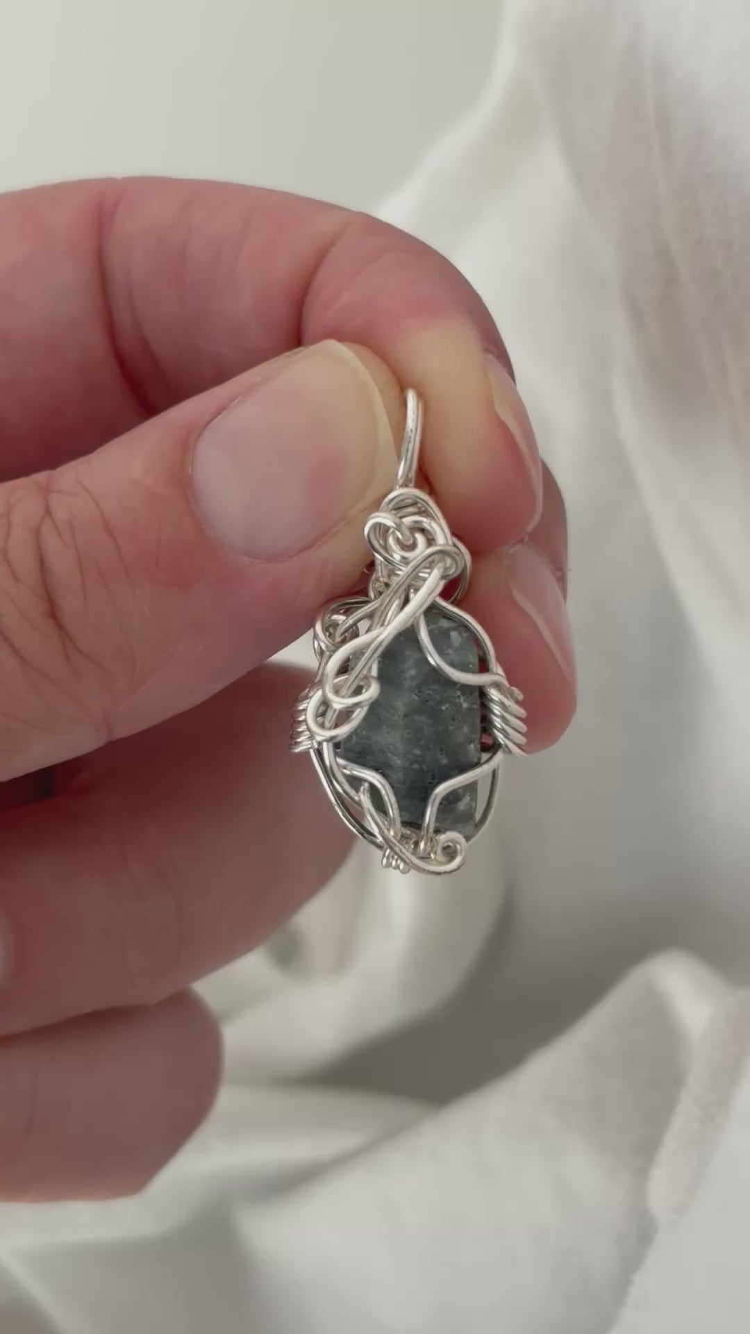 hand holding and moving the silver wire wrapped blue sapphire pendant around so all the sides of it are shown. done against a white background.